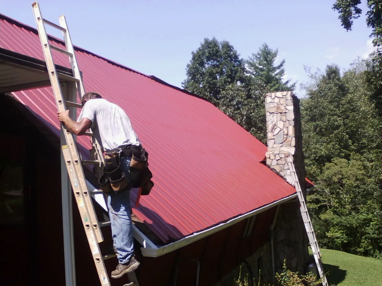 Roofer on a ladder beside a red metal roof and stone chimney — Soffit & Fascia Repair by Roofing Brothers in Putnam Valley